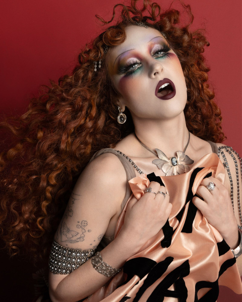 Chappell Roan appears in a MAC Viva Glam ad wearing a flowing costume and holding a flag. Her hair is styled in curls...