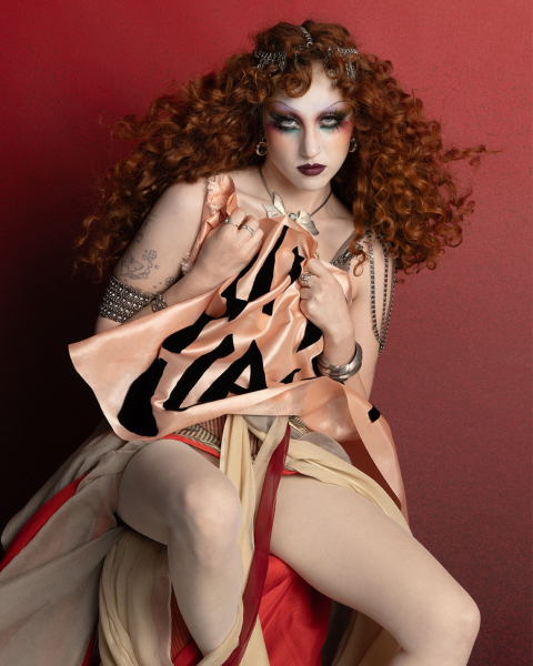 Chappell Roan appears in a MAC Viva Glam ad wearing a flowing costume and holding a flag. Her hair is styled in curls...